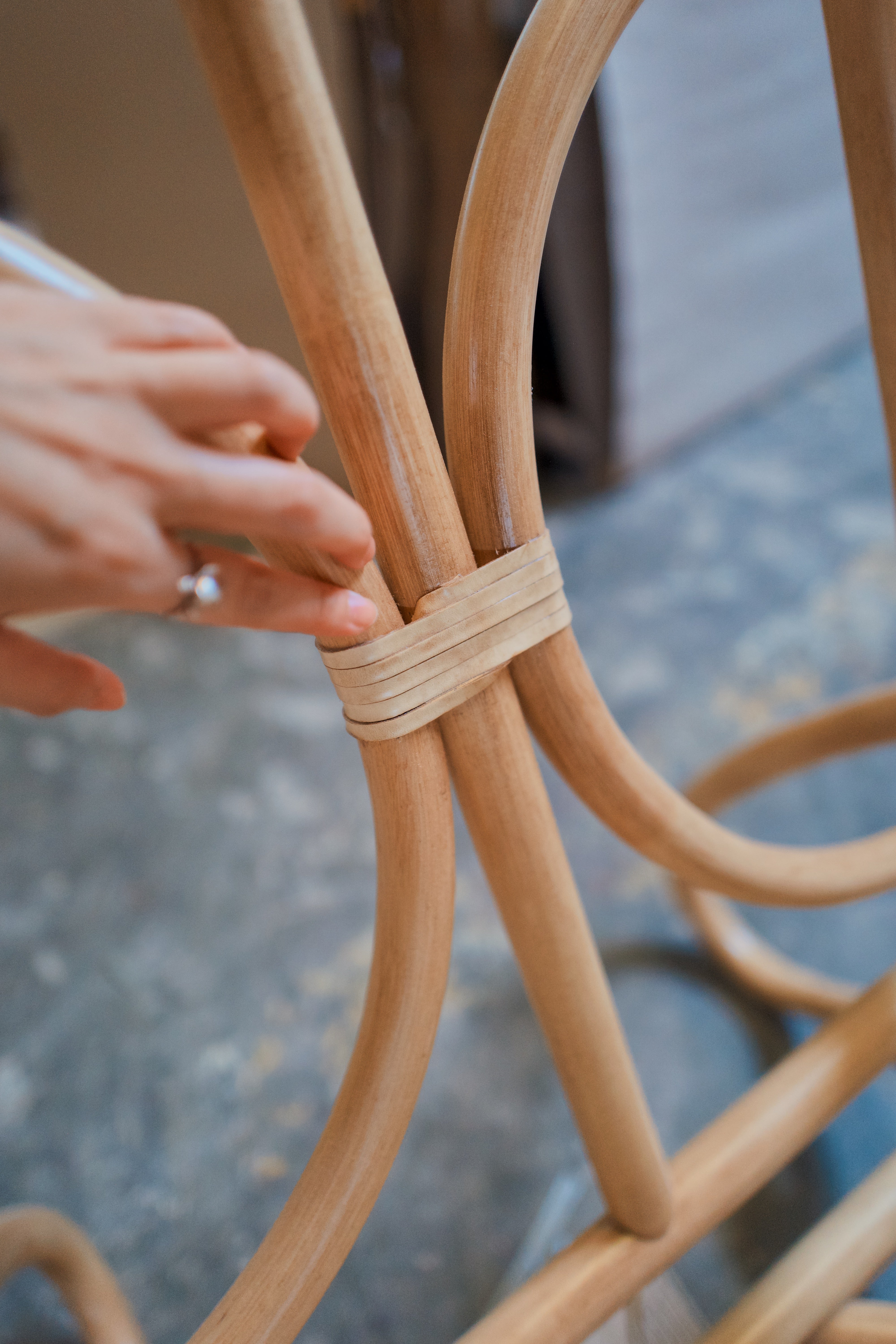 Close-up of a hand touching a light tan leather strip around a curved natural rattan chair frame, with a blurred indoor background.